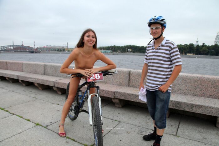 A thin tall girl Alexandra W takes off a T-shirt next to a cyclist at public