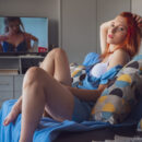 Redhead Elin Flame enjoys her popcorn while watching a film in the living room. She takes off her short dress and fingers her pussy.