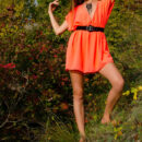 A smiling and confident Sofy frolics among the grass and plants in her bright orange dress like a carefree forest nymph.