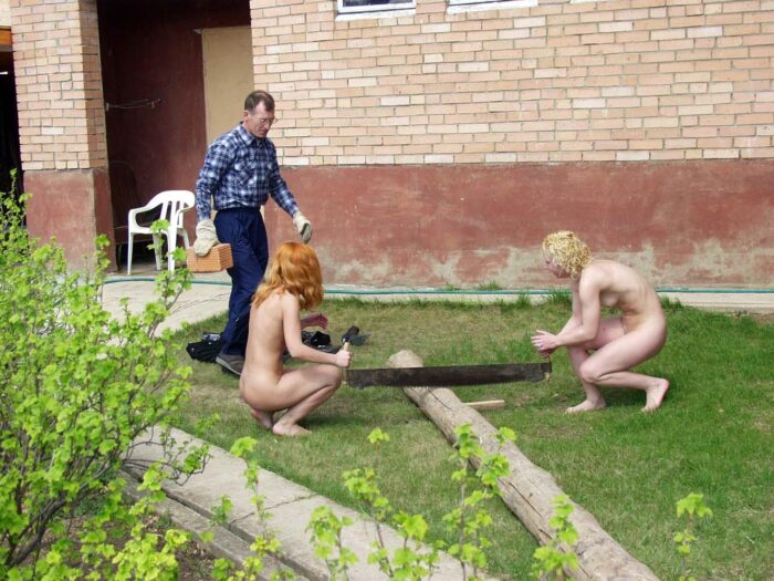 Two girls help a man saw a log naked